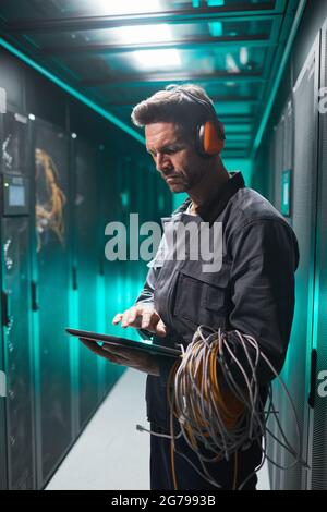 Vertical side view portrait of mature network engineer using digital tablet in server room during maintenance work in data center Stock Photo