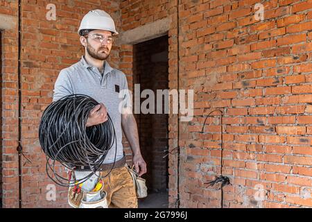 An electrician in a hard hat looks at the wall while holding an electric cable. Stock Photo