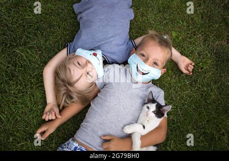 Two toddler boys lie on backs on green grass, looking up. Both wear blue medical protective masks with funny smiles. communication between children in Stock Photo