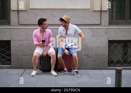 Two tourists sitting on their suitcases looking at each other and smiling. Stock Photo