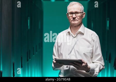 Waist up portrait of senior data scientist looking at camera and holding tablet while working with supercomputer in futuristic blue light, copy space Stock Photo
