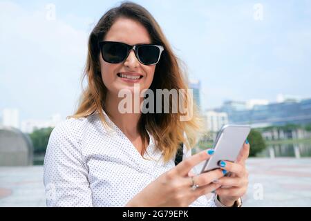Female entrepreneur in sunglasses standing outdoors, using application on smartphone and looking at camera Stock Photo