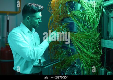 Side view portrait of handsome data engineer inspecting cables in server room while working with supercomputer in blue light, copy space Stock Photo