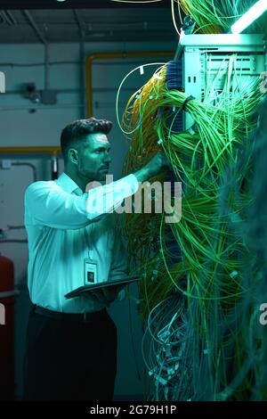 Vertical portrait of handsome data engineer inspecting cables in server room while working with supercomputer in blue light Stock Photo
