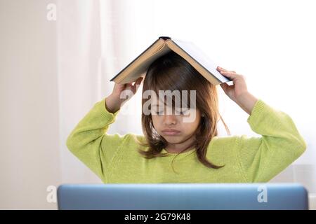 Sad elementary schooler girl studying at home with a book on her head because she is tired and angry - Little girl studying online and using internet Stock Photo