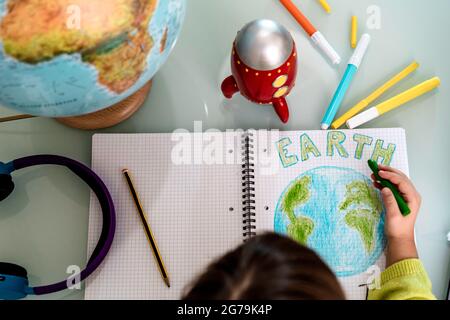 Top view of child girl draws planet earth with wax colors on school notebook for Earth day - Little activist girl writes the message Save the Planet - Stock Photo