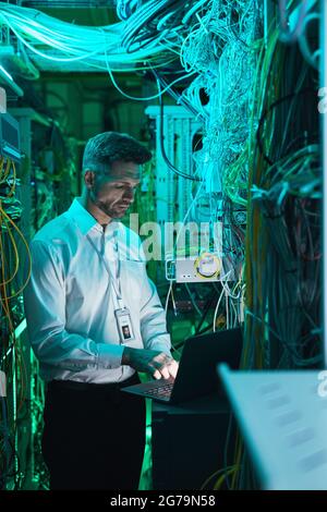 Vertical portrait of mature network technician using laptop in server room while working with supercomputer in data center Stock Photo