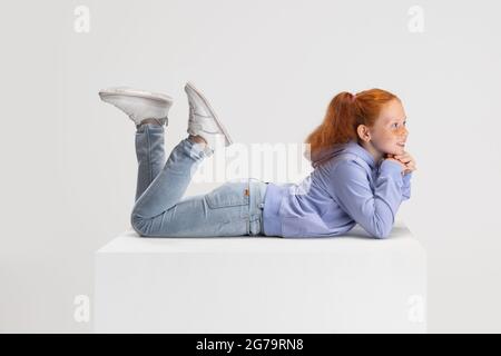 Side view. Portrait of smiling red-headed girl in casual clothes isolated on white studio background. Happy childhood concept. Sunny child. Looks Stock Photo