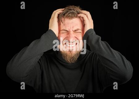 Exhausted bearded man portrait with headache, black background. Overworking, head pain concept. Young guy in black polo neck sweater. Exhausted and ti Stock Photo