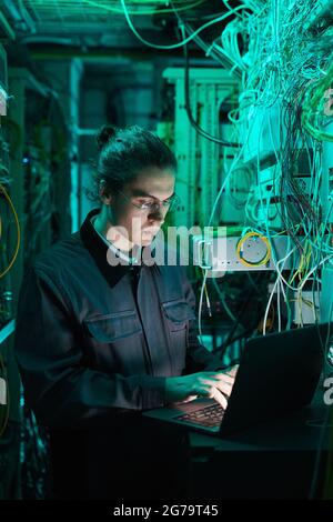 Vertical portrait of young network technician using laptop in server room while setting up supercomputer in data center Stock Photo