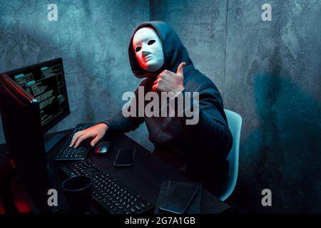 Anonymous hacker wearing face mask working on computer in dark room Stock Photo
