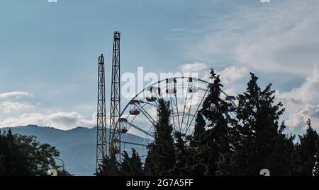 Silhouettes of a Ferris wheel, two cell towers, trees against the background of mountains and a beautiful cloudy sky in the amusement park of the city Stock Photo