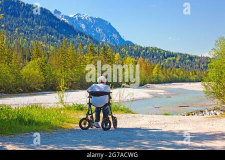 Old woman sits in a rollator on the banks of the Isar near Wallgau and enjoys the beautiful landscape Stock Photo