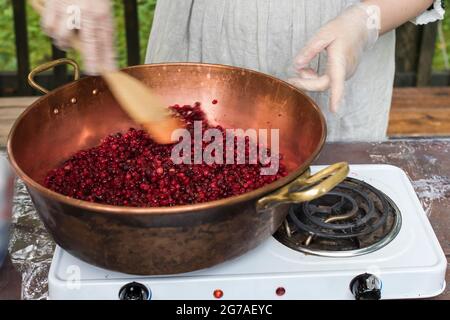 Cooking Finnish lingonberry jam with spices in a copper basin on a gas stove. Female hand with spoon Stock Photo