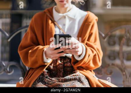 Young woman sitting on a bench with her cell phone. She is texting. Unrecognizable woman. Stock Photo