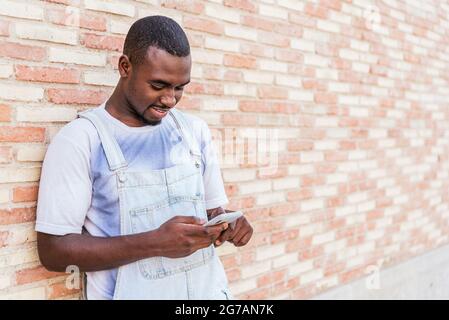 afro man checking his phone while leaning on wall. He dressed casual clothing ans smiles Stock Photo