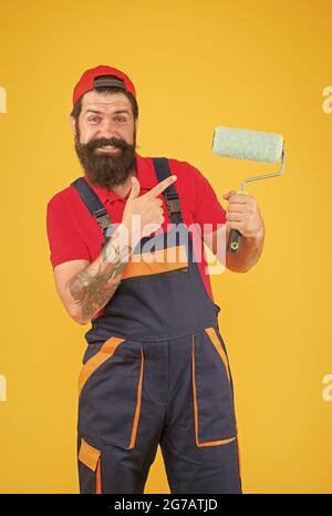 smiling bearded guy painter worker in boiler suit uniform pointing finger on paint roller tool for facing wall, repair Stock Photo