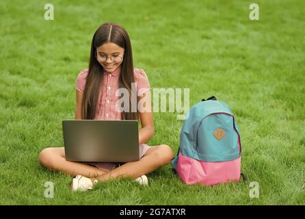 smart happy kid wear glasses using laptop for doing school homework online sitting in park on green grass with backpack, education Stock Photo