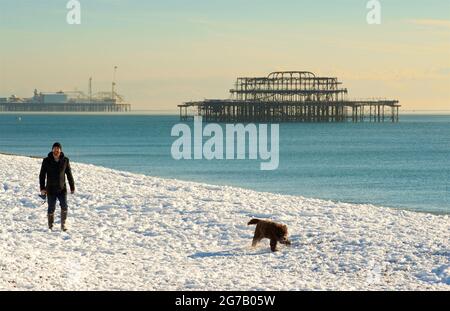 Snow on the beach, Brighton, England, with the Brighton Palace Pier and remains of the West pier behind. Man walking dog in the snow Stock Photo
