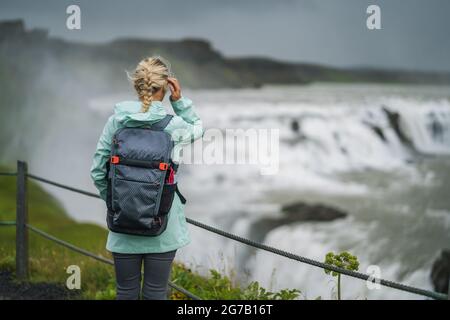 Blomd woman with backpack and green jacket visit Gullfoss powerful famous waterfall in Iceland. Famous place to visit on Golden Circle route of Iceland Stock Photo