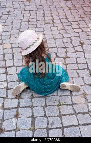 A girl in a straw hat sits on cobblestones Stock Photo