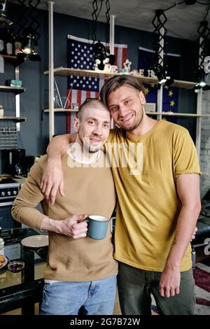 Portrait of happy hugging gay couple standing in living room and looking at camera Stock Photo