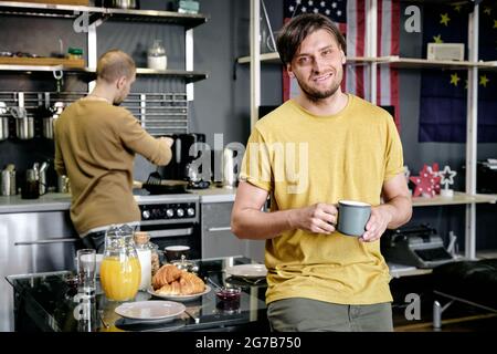 Smiling guy standing in kitchen and having drink, his husband cooking breakfast in background Stock Photo