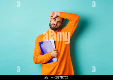 Education. Tired male student holding notebooks and wiping sweat off forehead, looking exhausted, standing in orange sweater against turquoise Stock Photo
