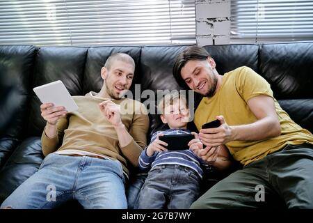 Joyful kid and his two fathers spending time together, they are sitting on sofa and playing games on mobile devices Stock Photo