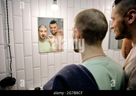 Happy young homosexual couple brushing teeth and looking at bathroom mirror Stock Photo