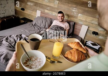 Young man bringing tray with breakfast to his handsome boyfriend in relaxing bed Stock Photo