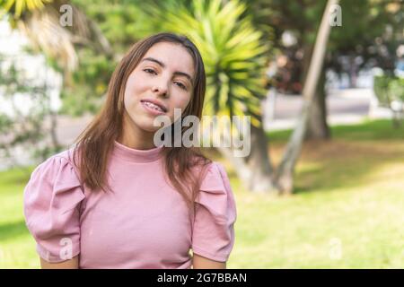 Portrait of a Latina teenage girl with braces looking at camera outdoors dressed in a pink blouse in a park Stock Photo