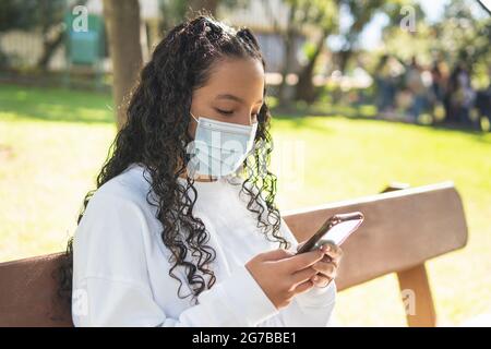 Teenage girl with curly hair sitting on a park bench sending text messages on her cell phone sitting on a bench Stock Photo