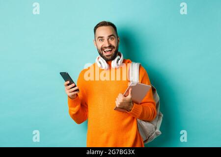 Handsome man student with headphones and backpack, holding digital tablet and smartphone, looking at camera, standing against turquoise background Stock Photo
