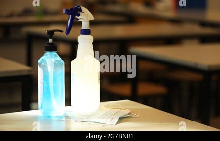 An empty school classroom with desks in the background. A face mask, bottle of hand sanitizer and cleaning spray sit in the sunlight in the foreground Stock Photo