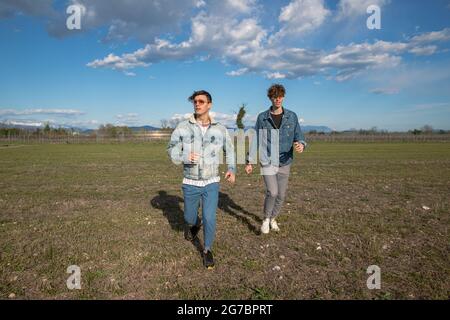 Two brothers run in a field of grass in the open air, young men dressed causally, wearing denim jackets. brotherhood concept. Stock Photo