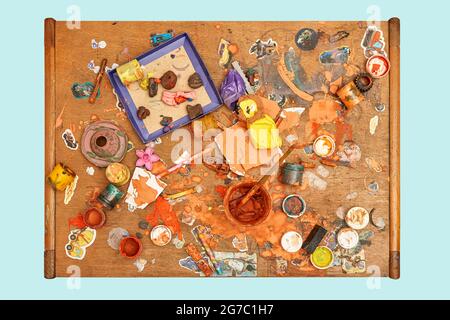 Jars of colorful paints brushes plasticine pieces and small toys on messy wooden table in light children room upper view isolated on blue background Stock Photo
