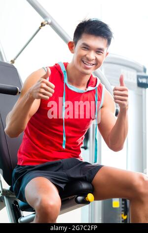 Asian Chinese man having fitness training or workout in gym doing sport to build up muscle on a weight machine Stock Photo