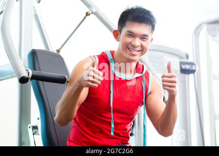 Asian Chinese man having fitness training or workout in gym doing sport to build up muscle on a weight machine Stock Photo