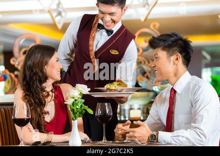 Asian Chinese couple - Man and woman - or lovers having a date or romantic dinner in a fancy restaurant while the waiter is serving food Stock Photo