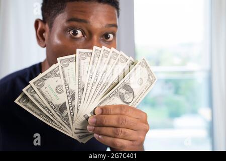 Close up of African American man holds up us currency to his face with eyes wide open, camera focuses on the money. Stock Photo