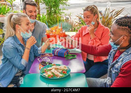 Friends drinking coktail and eating snack tapas in a restaurant bar outside in summer days with face mask on to be protected from coronavirus - Happy Stock Photo