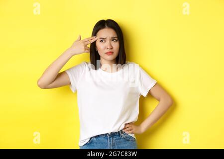 Woman showing kill me please gesture, shooting herself in head with finger gun from boredom, standing upset over yellow background Stock Photo