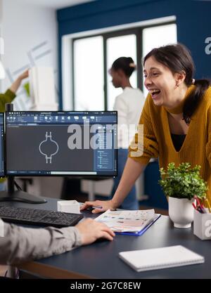 Excited mechanical engineers laughing and discussing about manufacturing new project, taking break. Man technician working at pc, screen shows 3D engine made with CAD software, using computer Stock Photo