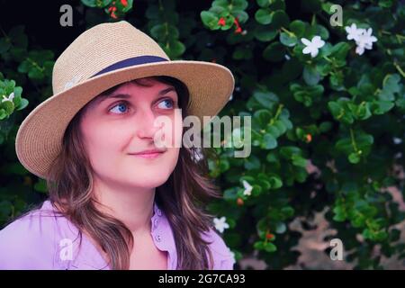 Portrait of a young woman in a straw hat on a background of flowers, copy space. The woman in the red dress smiles and holds her hat in her hand. Stock Photo