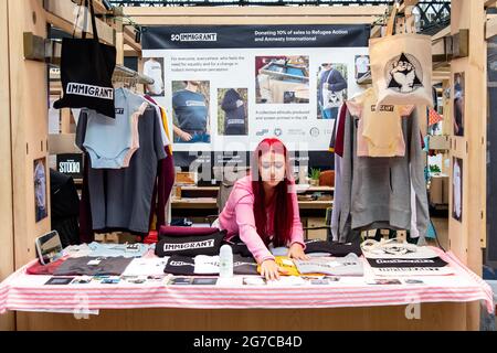 London- July, 2021: A girl selling a line of fashion items at Spitalfields market in East London with the word / branding  ‘Immigrant’ Stock Photo