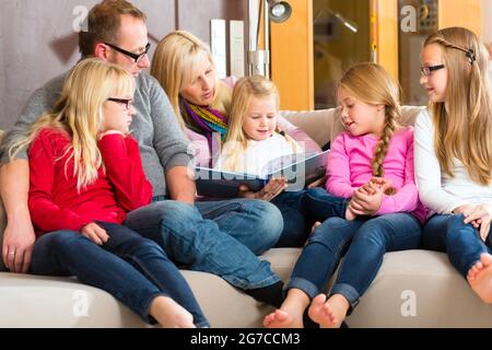 Family sitting together with mother, father and children comfortable on sofa, listening to fairy tale from story book Stock Photo