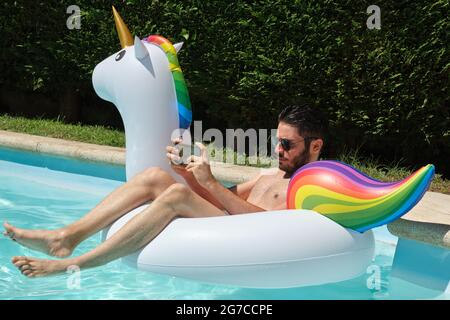 Young man wearing sunglasses playing on the phone on a unicorn inflatable ring in a swimming pool. Summer concept. Stock Photo