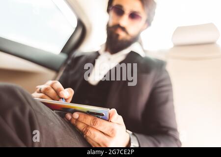 Stylish handsome Hindu businessman with glasses and a beard works in the backseat of the car and uses the phone. Safe and comfortable luxury travel Stock Photo