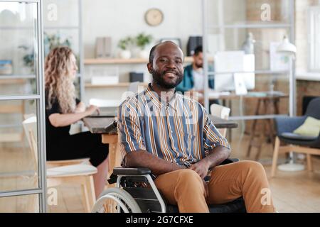 Portrait of African young businessman sitting on wheelchair and smiling at camera while working at office with his colleagues Stock Photo